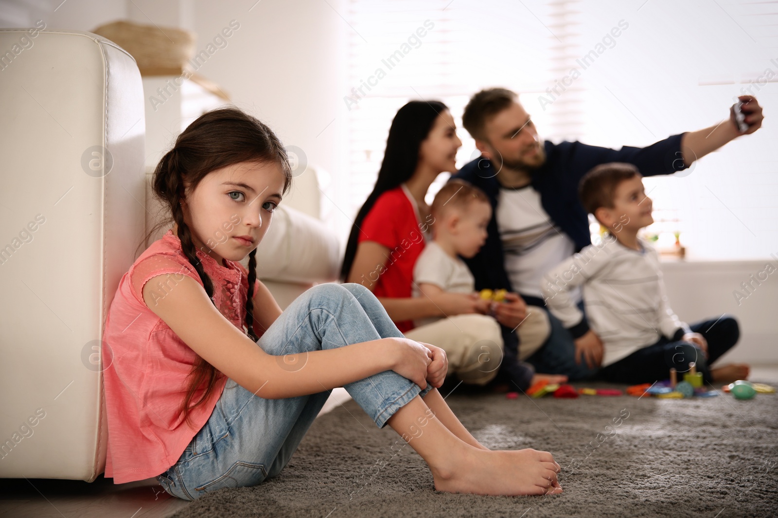 Photo of Unhappy little girl feeling jealous while parents taking selfie with other children at home