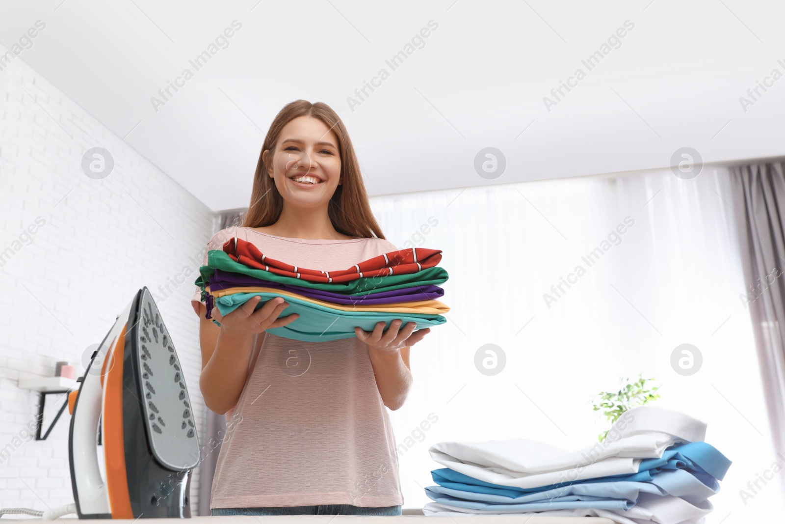 Photo of Young woman with folded clothes near ironing board at home. Space for text