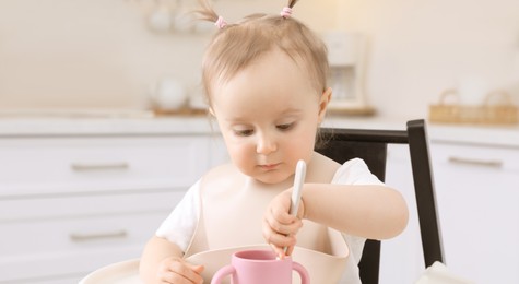 Image of Cute little baby with spoon and cup in high chair indoors