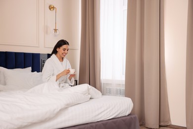 Photo of Happy young woman wearing bathrobe drinking coffee on bed in hotel room