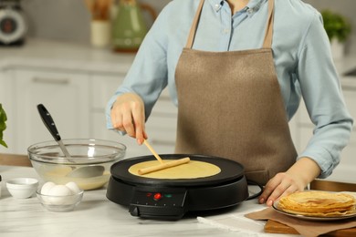 Woman cooking delicious crepe on electric maker at white marble table in kitchen, closeup