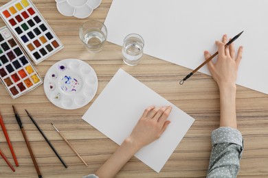 Woman painting with watercolor on blank paper at wooden table, top view