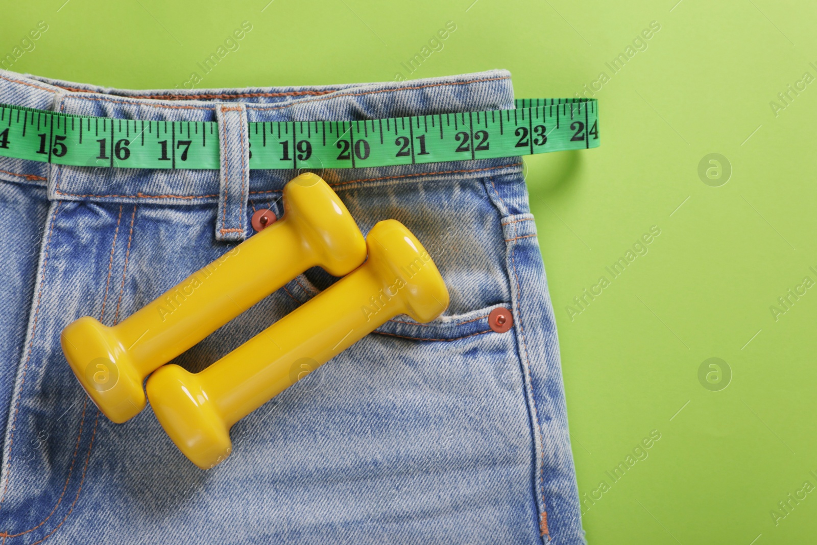 Photo of Jeans, dumbbells and measuring tape on light green background, flat lay. Weight loss concept