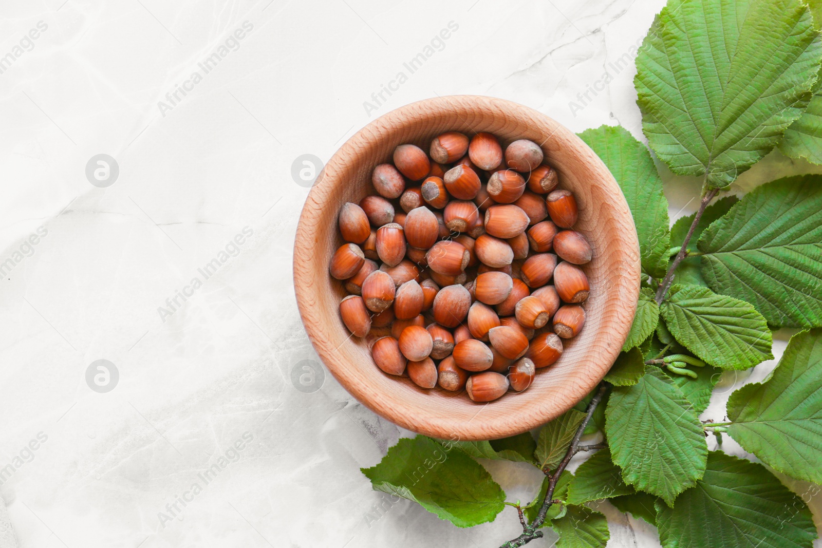 Photo of Tasty hazelnuts in wooden bowl and branch on light marble table, flat lay. Space for text