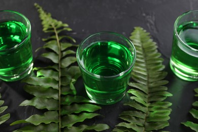 Photo of Absinthe in shot glasses and green leaves on black table, closeup. Alcoholic drink