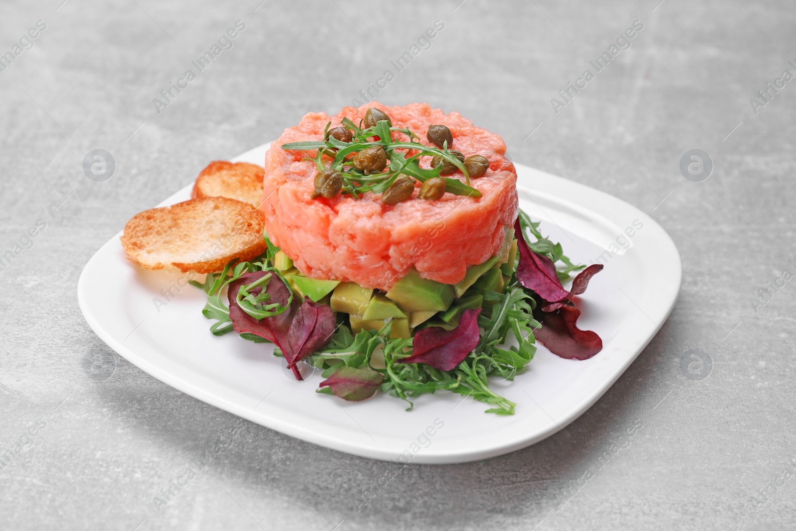 Photo of Tasty salmon tartare with avocado, greens and croutons on grey table