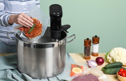 Photo of Woman putting vacuum packed meat into pot with sous vide cooker at wooden table, closeup. Thermal immersion circulator