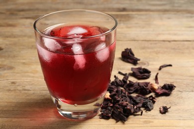 Glass of delicious iced hibiscus tea and dry flowers on wooden table, closeup
