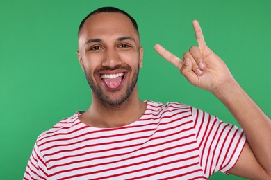 Photo of Happy young man showing his tongue and rock gesture on green background