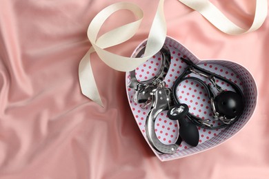 Photo of Heart shaped gift box with different sex toys and ribbon on pink silk fabric, flat lay. Space for text