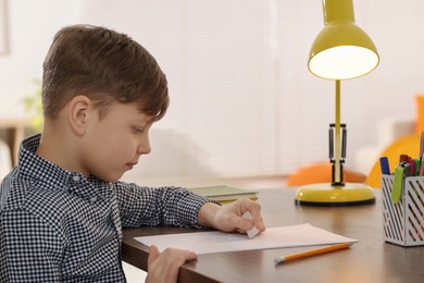 Little boy erasing mistake in his notebook at wooden desk indoors