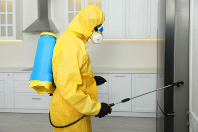 Photo of Pest control worker in protective suit spraying insecticide near refrigerator indoors