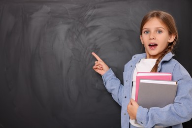 Photo of Cute schoolgirl with books pointing at something on blackboard. Space for text