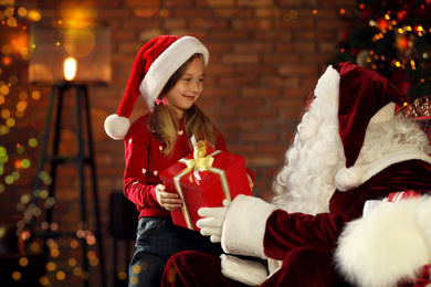 Photo of Santa Claus giving Christmas gift to little girl indoors