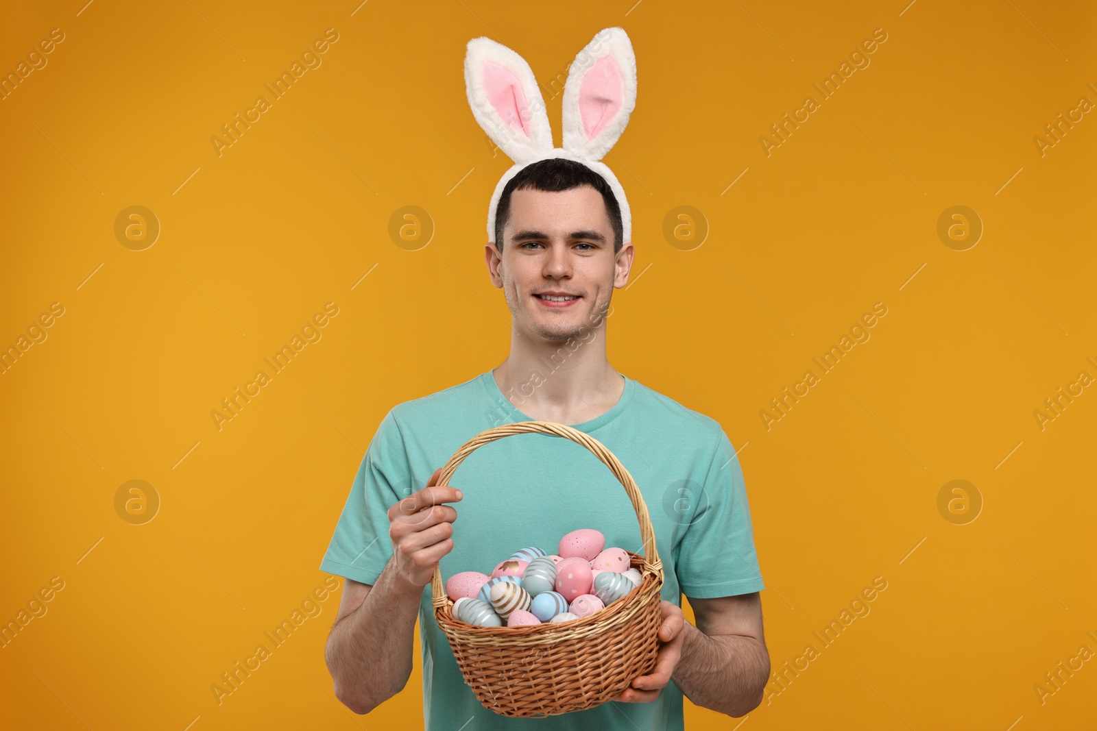 Photo of Easter celebration. Handsome young man with bunny ears holding basket of painted eggs on orange background