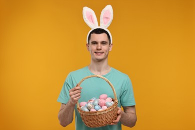 Photo of Easter celebration. Handsome young man with bunny ears holding basket of painted eggs on orange background