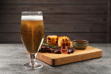 Photo of Glass of beer, delicious fried steak, corn and sauce on grey table. Space for text