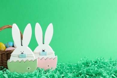 Photo of Wooden bunnies with protective masks and painted eggs in basket on green background, space for text. Easter holiday during COVID-19 quarantine