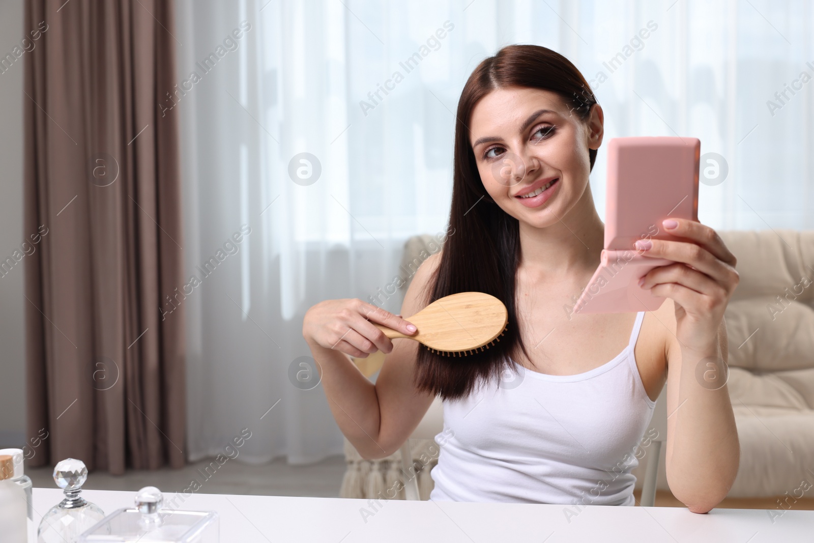 Photo of Beautiful woman brushing her hair and looking into compact mirror in room, space for text