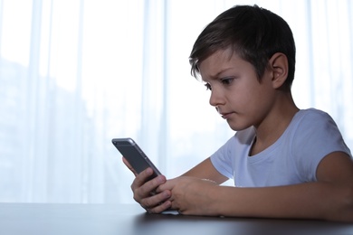 Photo of Terrified child with smartphone at table indoors. Danger of internet