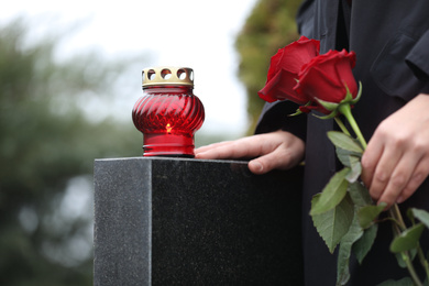 Photo of Woman holding red roses near black granite tombstone with candle outdoors, closeup. Funeral ceremony