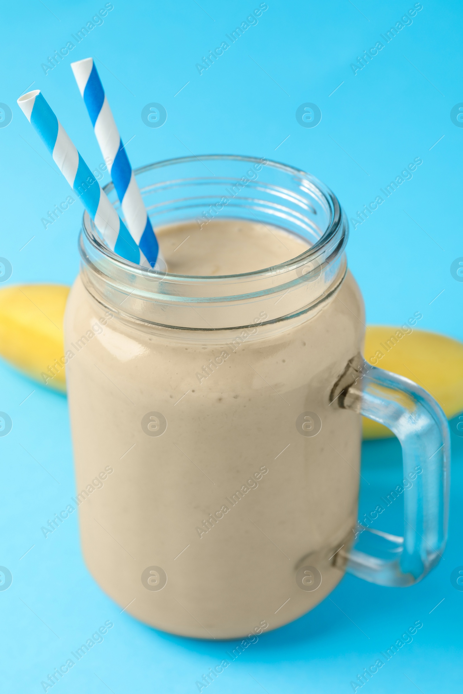 Photo of Mason jar of tasty banana smoothie with straws and fresh fruit on light blue background