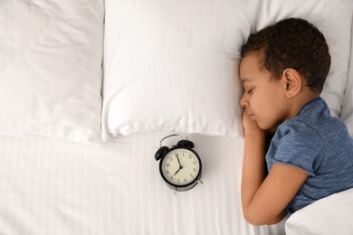 Photo of Cute little African-American boy with alarm clock sleeping in bed, top view. Space for text