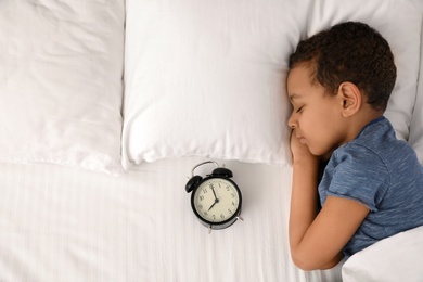 Cute little African-American boy with alarm clock sleeping in bed, top view. Space for text