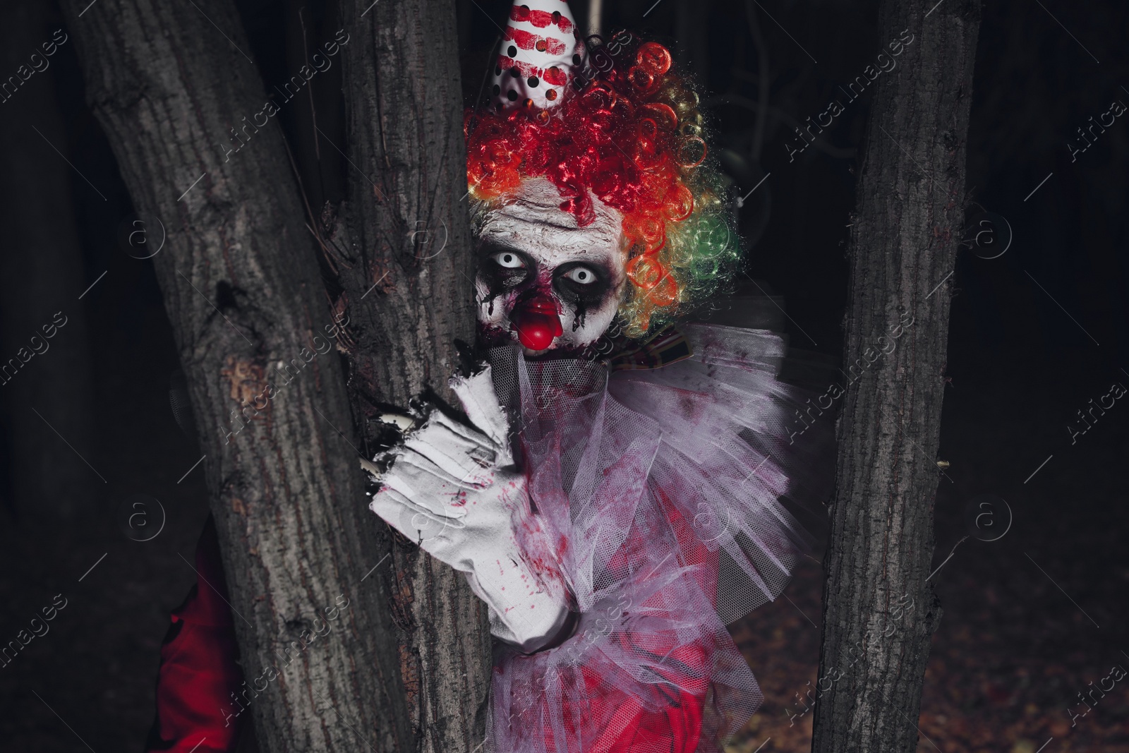 Photo of Terrifying clown hiding behind trees outdoors at night. Halloween party costume