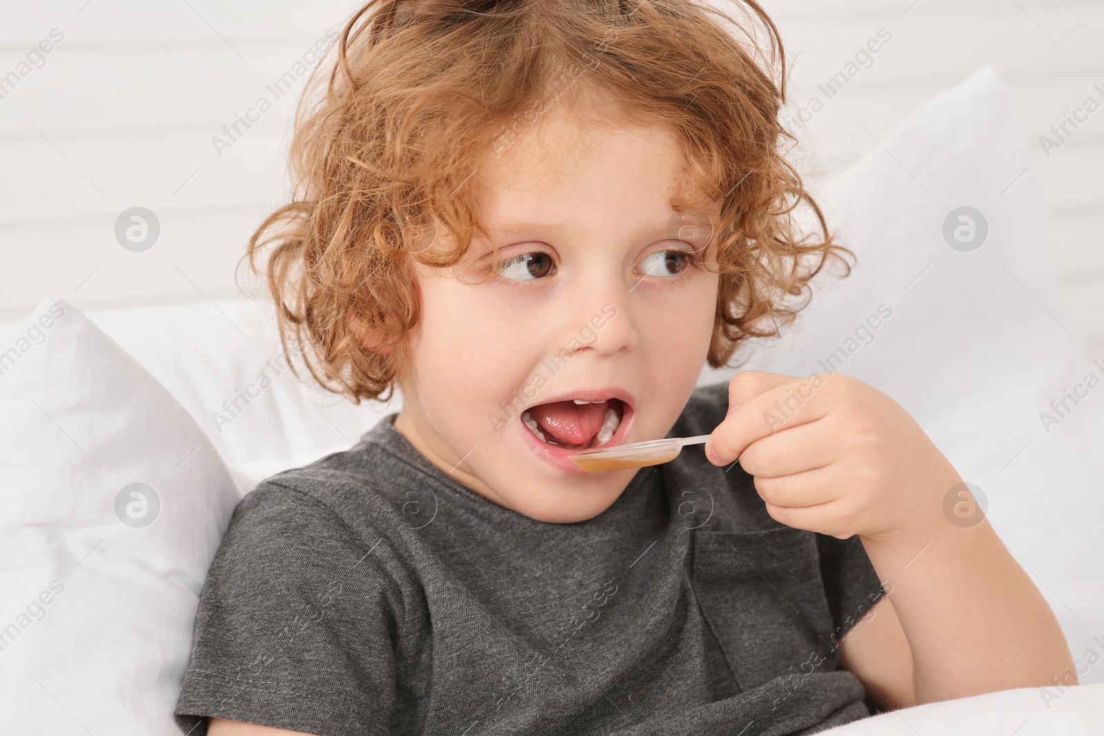 Photo of Cute boy taking measuring spoon with cough syrup on bed. Effective medicine