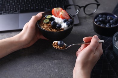 Woman eating tasty granola with yogurt and berries at workplace, closeup