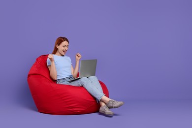 Photo of Happy young woman with laptop sitting on beanbag chair against lilac background, space for text