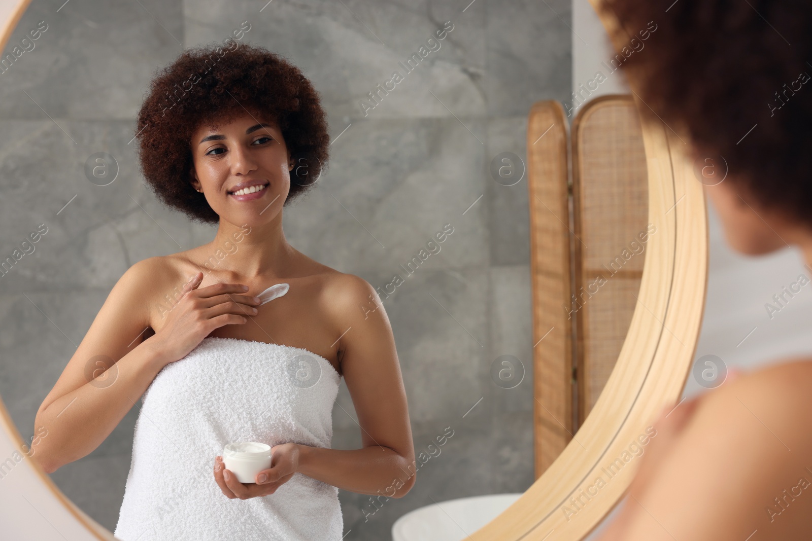 Photo of Beautiful young woman applying cream onto body in bathroom