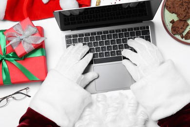 Photo of Santa Claus using laptop, closeup. Gift boxes, eyeglasses and biscuits on white table, top view