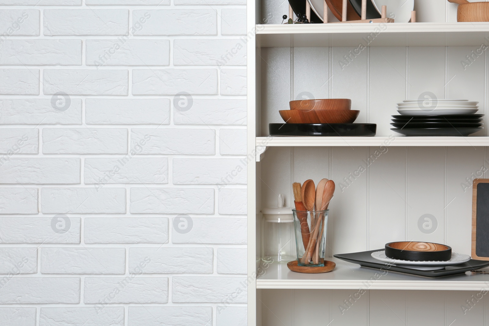 Photo of White shelving unit with set of dishware near brick wall. Space for text