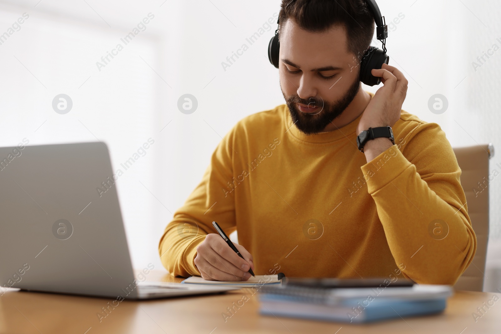 Photo of Young man in headphones writing down notes during webinar at table in room