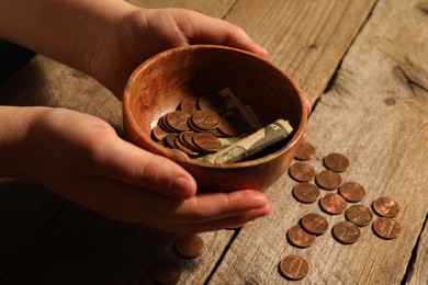 Donate and give concept. Woman holding bowl with coins and dollar banknotes at wooden table, closeup