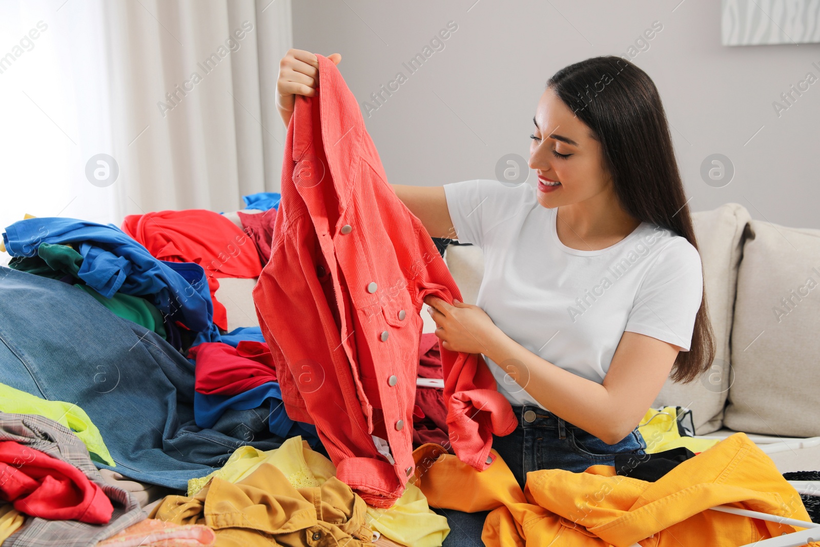 Photo of Happy young woman with lots of clothes on sofa in room. Fast fashion