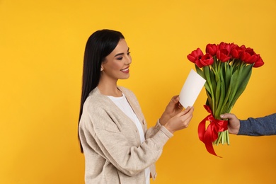 Photo of Happy woman receiving red tulip bouquet and greeting card from man on yellow background. 8th of March celebration