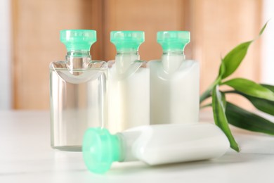 Photo of Mini bottles of cosmetic products and green branch on white table against blurred background, closeup