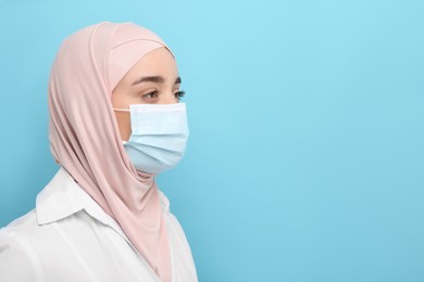 Photo of Muslim woman wearing hijab, medical uniform and protective mask on light blue background, space for text