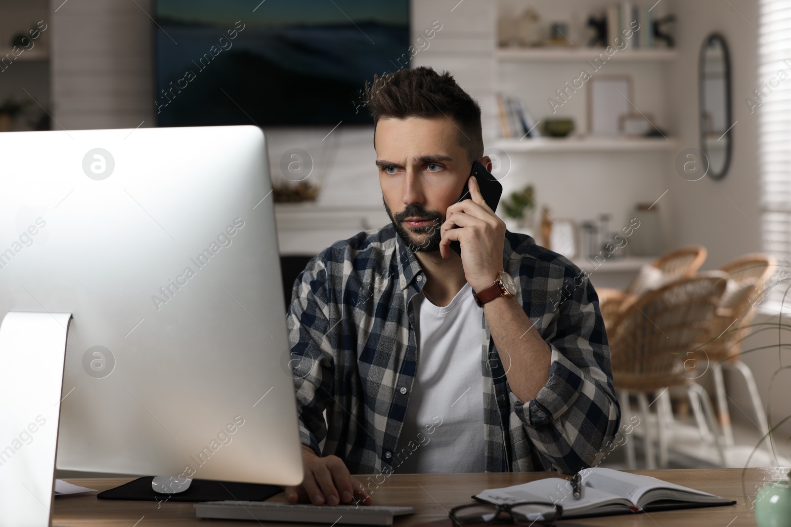 Photo of Man talking on phone while working with computer at table in home office