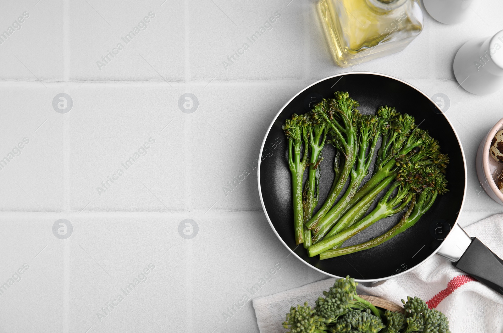 Photo of Frying pan with tasty cooked broccolini and other products on white tiled table, flat lay. Space for text