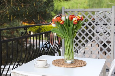 Photo of Beautiful colorful tulips in glass vase and cup of drink on white table at balcony