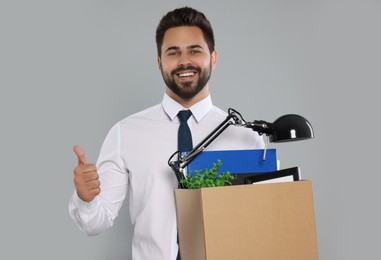 Happy unemployed man with box of personal office belongings showing thumb up on light grey background