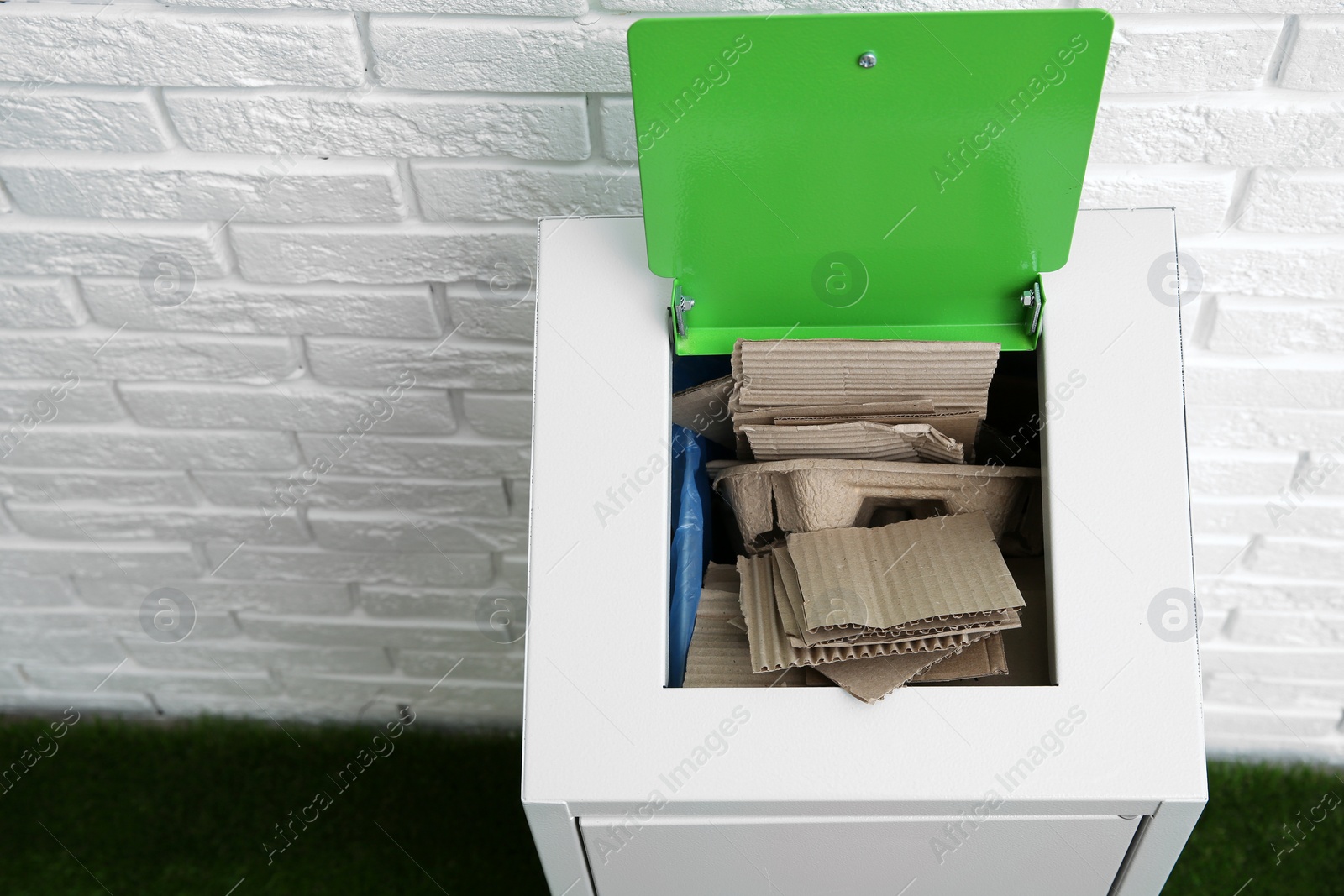 Photo of Trash bin with cardboard near brick wall indoors, space for text. Recycling concept