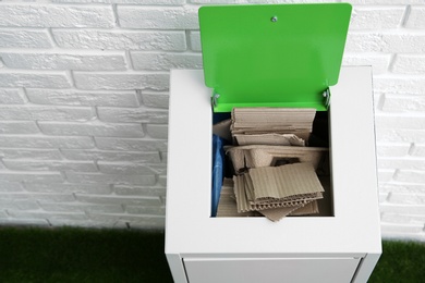 Trash bin with cardboard near brick wall indoors, space for text. Recycling concept