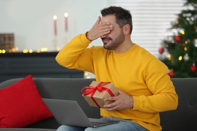 Celebrating Christmas online with exchanged by mail presents. Man covering eyes before opening gift box during video call on laptop at home