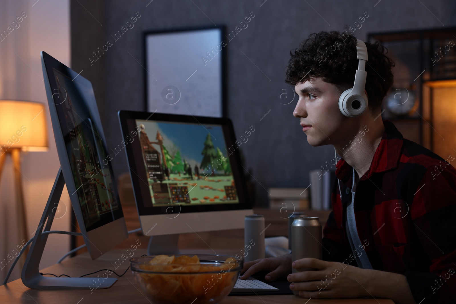 Photo of Young man with energy drink and headphones playing video game at wooden desk indoors