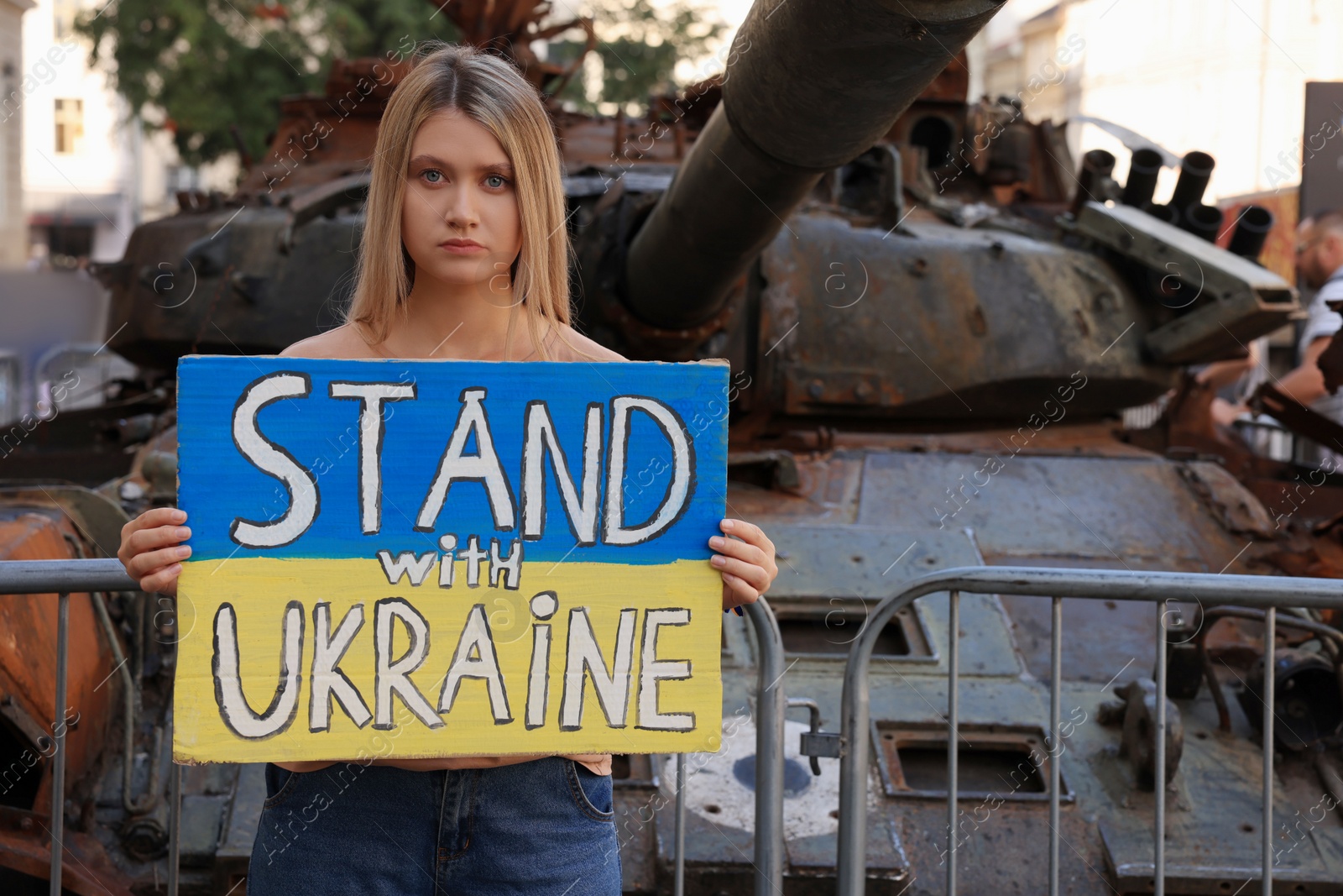 Photo of Sad woman holding poster in colors of national flag with words Stand with Ukraine near broken tank on city street